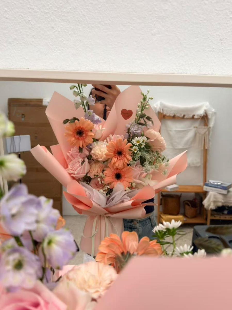 Vibrant Girl - Fresh and Sweet Hand-Tied Bouquet in Light Pink Packaging Featuring Peach-Pink Gerberas, Light Pink Roses, Purple Florals, and White Daisies